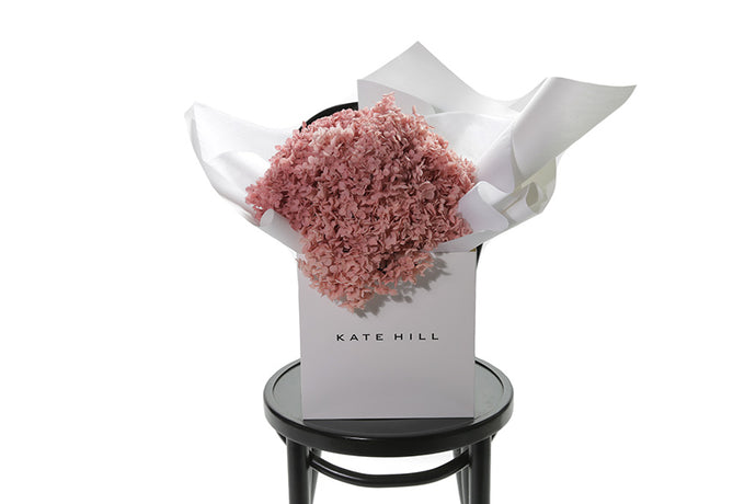 A bouquet of soft pastel pink preserved dried Hydrangeas, beautifully wrapped in the Kate Hill Flowers signature presentation. Bouquet presented in Kate Hill flower bag, sitting on black bentwood chair with white background.