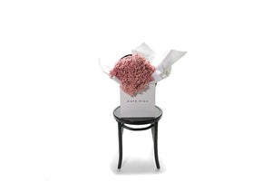 Wide image of pink preserved Hydrangea bouquet to give an idea of size. A bouquet of soft pastel pink preserved dried Hydrangeas, beautifully wrapped in the Kate Hill Flowers signature presentation. Bouquet presented in Kate Hill flower bag, sitting on black bentwood chair with white background.