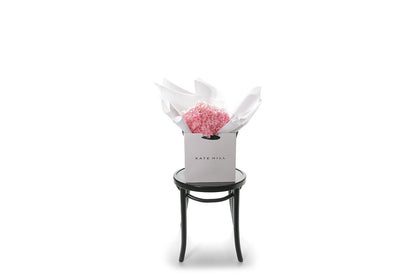 Wide image of a bouquet of candy pink bouquet of preserved dried hydrangeas, beautifully presented in signature white wrapping and flower bag. Flower bouquet sitting on a black bentwood chair with white wall background.