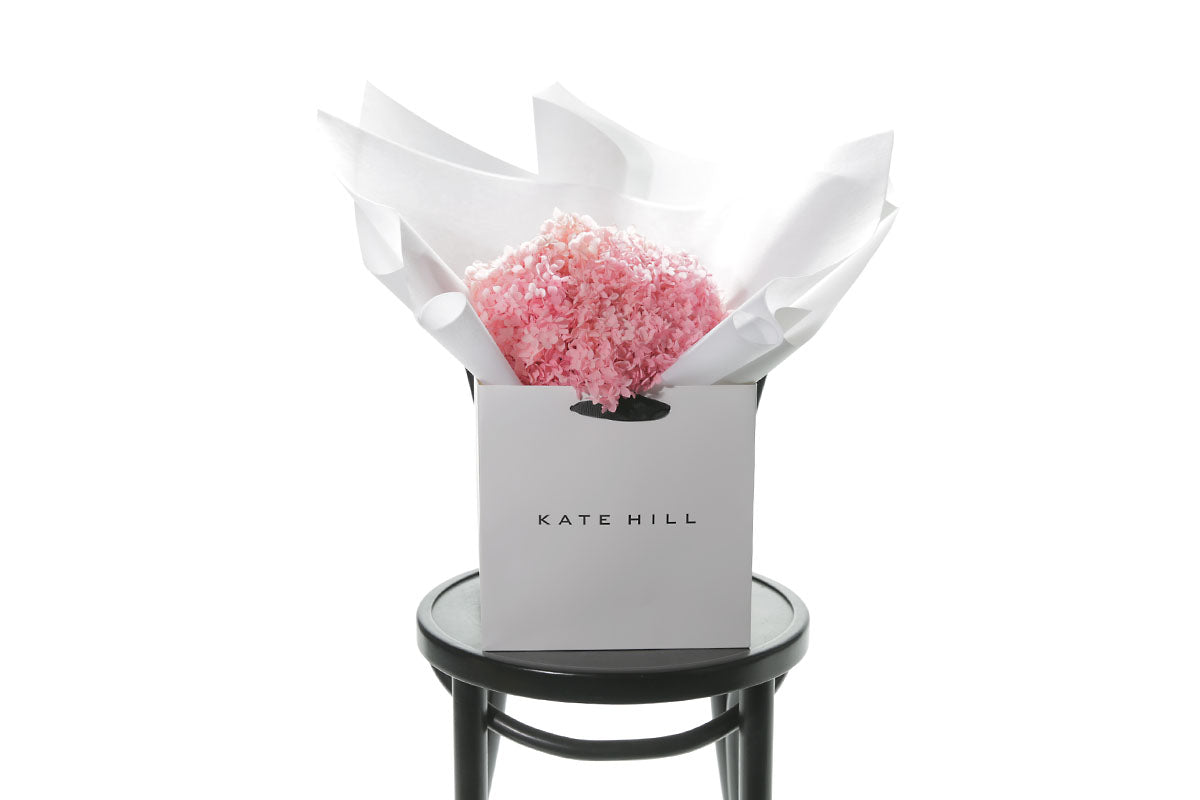 A bouquet of candy pink bouquet of preserved dried hydrangeas, beautifully presented in signature white wrapping and flower bag. Flower bouquet sitting on a black bentwood chair with white wall background.