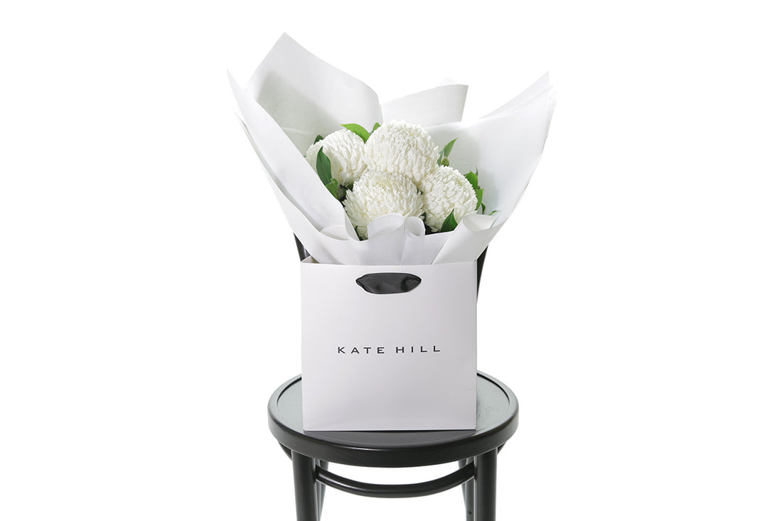 A small posy flower bouquet of white disbuds and a hint of foliage. Bouquet wrapped in beautiful white signature wrapping in flower bag. Bouquet bag sitting on a black bentwood chair with white wall background.