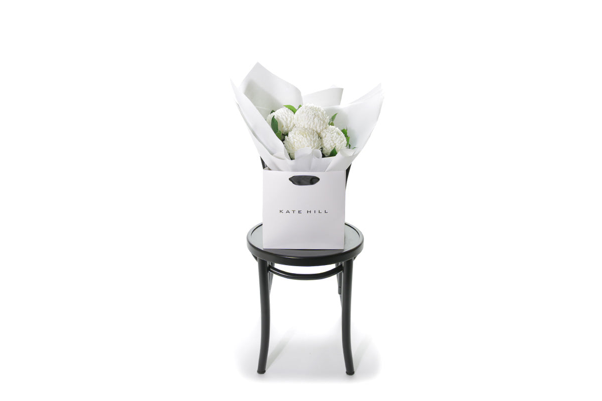 Wide image of small posy flower bouquet of white disbuds and a hint of foliage. Bouquet wrapped in beautiful white signature wrapping in flower bag. Bouquet bag sitting on a black bentwood chair with white wall background.