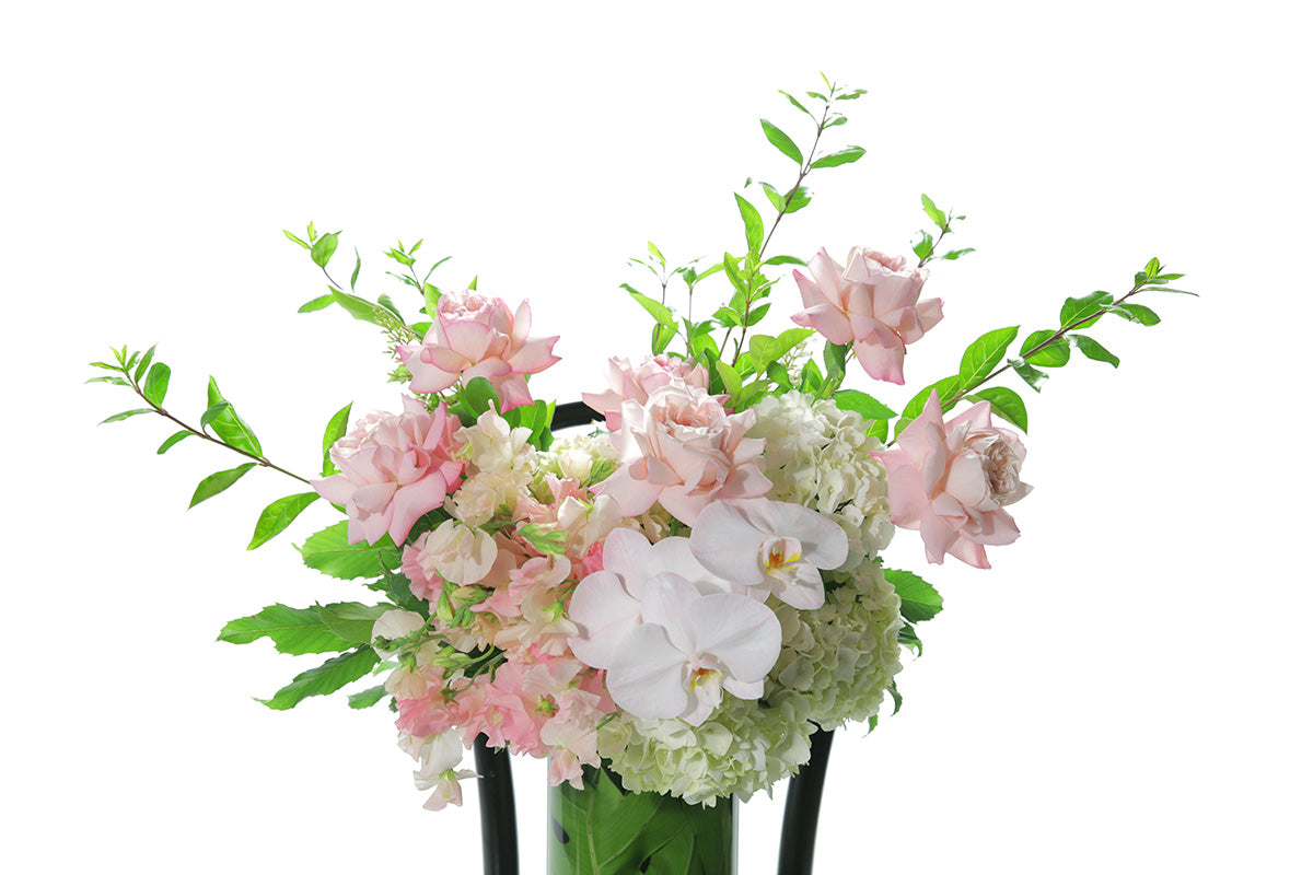Close up image of large vase design. A 25cm cylinder tall vase design, featuring mixed soft pink and mid pink seasonal flowers. Tall cylinder vase design sitting on a black bentwood chair with white background. Flowers sitting on chair with white wall background.