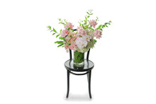 Wide image of Portia vase design. A 25cm cylinder tall vase design, featuring mixed soft pink and mid pink seasonal flowers. Tall cylinder vase design sitting on a black bentwood chair with white background. Flowers sitting on chair with white wall background.