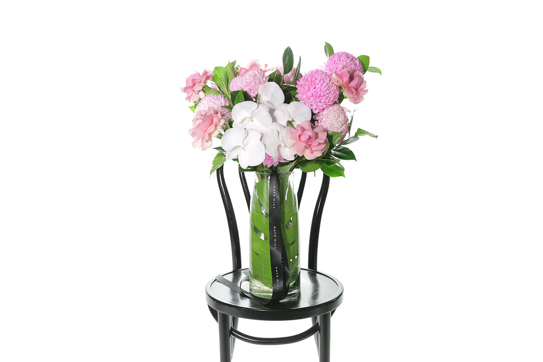 Kate Hill Flowers – large soft-pink celebration vase with roses, chrysanthemums and orchids in a monstera-lined glass.
