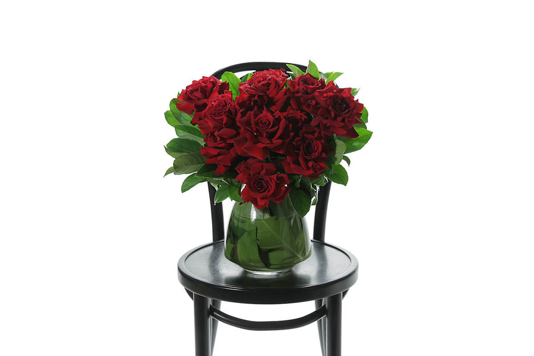 A green leaf lined tapered vase featuring 10 stems of red roses and green foliage, sitting on a black bentwood chair in front of a white wall.