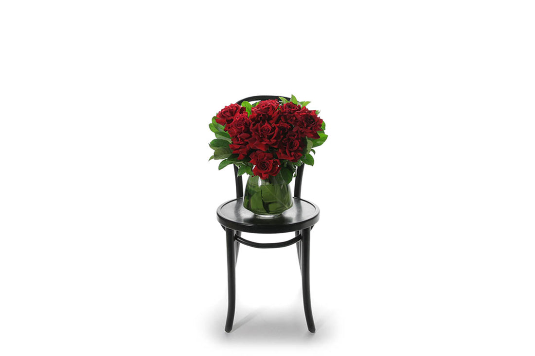 Wide image of a green leaf lined tapered vase featuring 10 stems of red roses and green foliage, sitting on a black bentwood chair in front of a white wall.