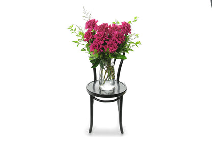 Hot pink reflexed roses displayed in a clear glass tapered vase sitting on a black bentwood chair with white background. Wide image of vase design.
