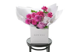 Large hot pink, mid pink and blush seasonal bouquet wrapped in signature Kate Hill flowers presentation. Bouquet is placed into flower bag to ensure the bouquet remains fresh in water. Bouquet bag is sitting on a black bentwood chair with white background.