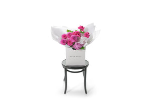 Wide image of Scarlett bouquet on black bentwood chair. Large hot pink, mid pink and blush seasonal bouquet wrapped in signature Kate Hill flowers presentation. Bouquet is placed into flower bag to ensure the bouquet remains fresh in water. Bouquet bag is sitting on a black bentwood chair with white background.