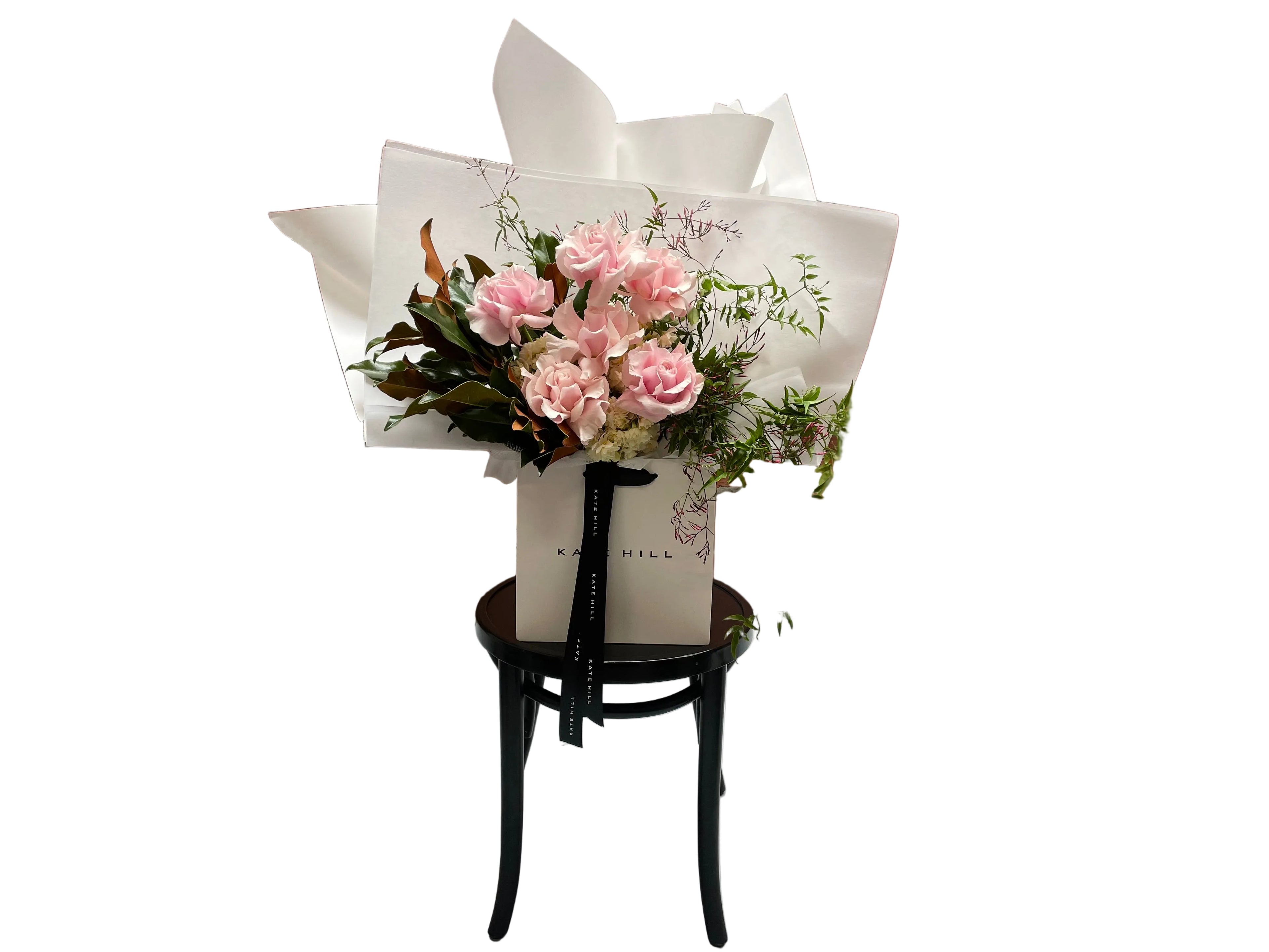 A soft pastel blush pink, cream and jasmine flower bouquet, wrapped in signature Kate Hill style and sitting on a black bentwood chair in front of white wall.