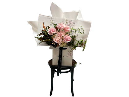 A soft pastel blush pink, cream and jasmine flower bouquet, wrapped in signature Kate Hill style and sitting on a black bentwood chair in front of white wall.