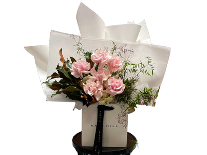 Closer image of a soft pastel blush pink, cream and jasmine flower bouquet, wrapped in signature Kate Hill style and sitting on a black bentwood chair in front of white wall.