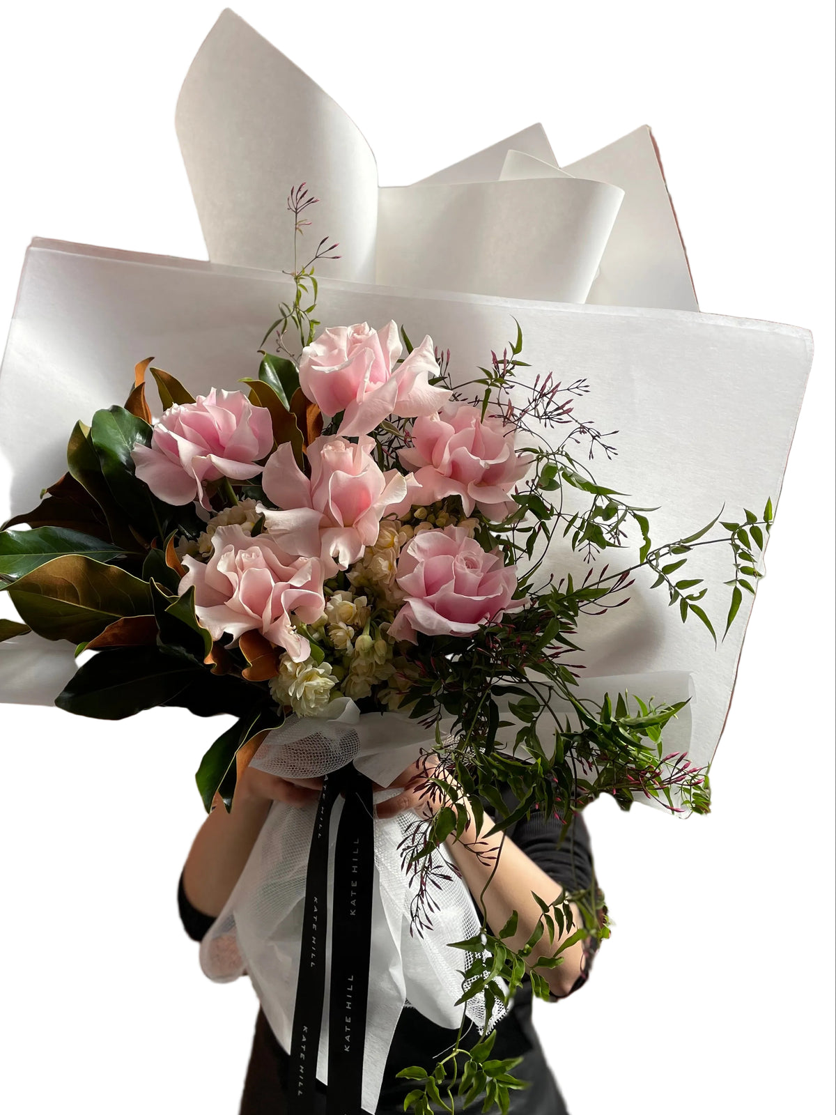 A soft pastel blush pink, cream and jasmine flower bouquet, wrapped in signature Kate Hill style and florist holding bouquet in front of white wall.