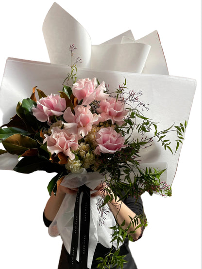 A soft pastel blush pink, cream and jasmine flower bouquet, wrapped in signature Kate Hill style and florist holding bouquet in front of white wall.