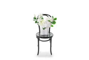 White ceramic wave vase displaying white and green seasonal flowers and foliages. Sia wave vase sitting on black bentwood chair with white background. Wide image of the white wave vase design on chair.