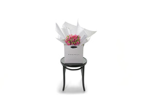 Small dome posy of fragrant pink sweet-peas, gift wrapped beautifully in white signature wrapping. Bouquet placed into Kate Hill Flower Bag and is sitting on a black bentwood chair with white background. Wide image of the bouquet on chair.