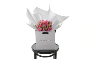 Small dome posy of fragrant pink sweet-peas, gift wrapped beautifully in white signature wrapping. Bouquet placed into Kate Hill Flower Bag and is sitting on a black bentwood chair with white background.