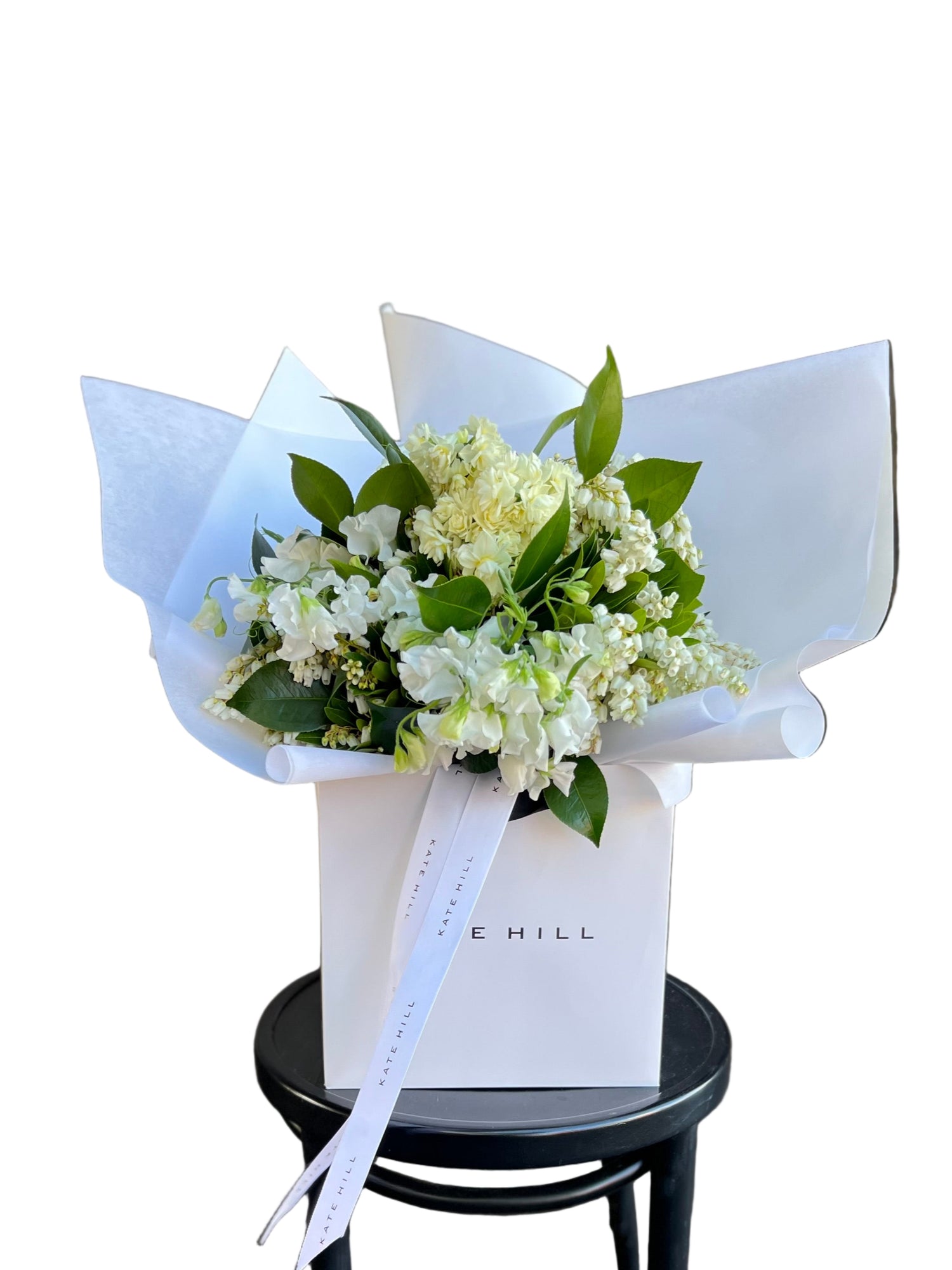 Bouquet of white, cream and lemon Spring flowers in a white Kate Hill Flowers bag, sitting on  black bentwood chair against a white wall.
