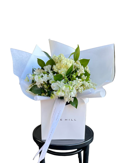 Bouquet of white, cream and lemon Spring flowers in a white Kate Hill Flowers bag, sitting on  black bentwood chair against a white wall.