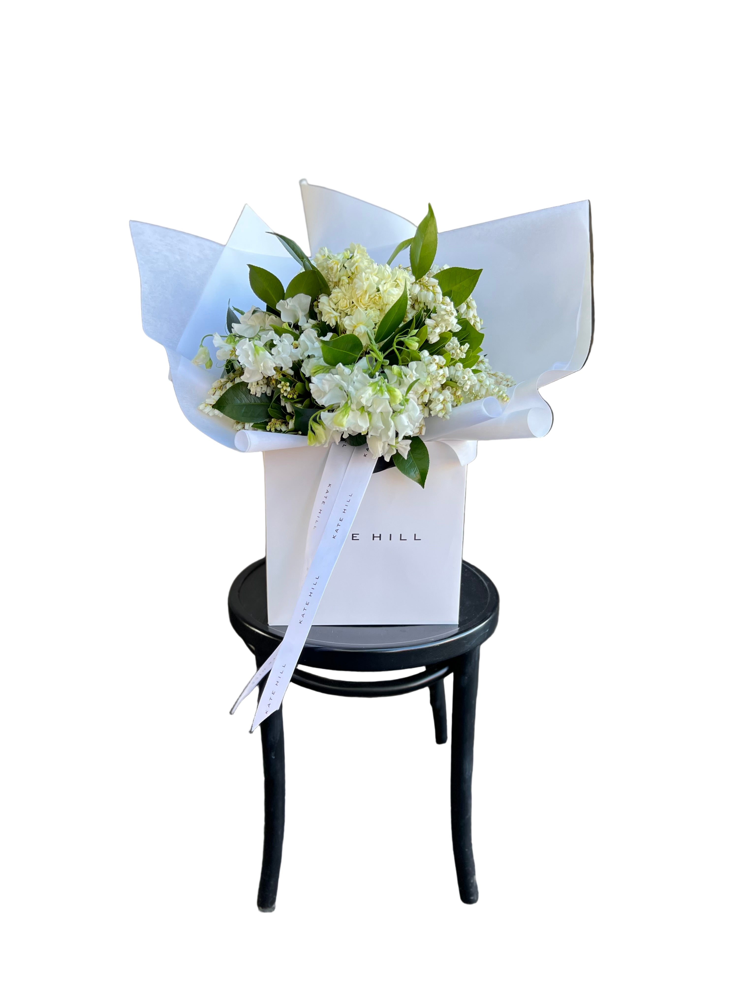 Bouquet of white, cream and lemon Spring flowers in a white Kate Hill Flowers bag, sitting on  black bentwood chair against a white wall.