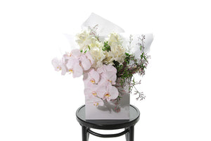 Large bouquet of white roses, blush phalaenopsis orchids and foliage. Wrapped in white signature wrapping, displayed into a kate hill flower bag and sitting on a black bentwood chair with white background.