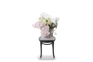Wide image of Stella large bouquet. Large bouquet of white roses, blush phalaenopsis orchids and foliage. Wrapped in white signature wrapping, displayed into a kate hill flower bag and sitting on a black bentwood chair with white background.