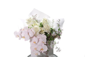 Close up image of Stella. Large bouquet of white roses, blush phalaenopsis orchids and foliage. Wrapped in white signature wrapping, displayed into a kate hill flower bag and sitting on a black bentwood chair with white background.