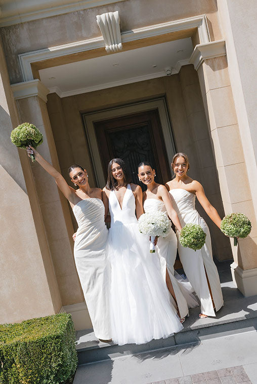 Bride and bridesmaids holding wedding flowers at Melbourne wedding in white dresses standing together in front of the entrance to a wedding venue