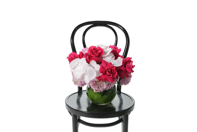 A pretty hot pink and blush pastel pink vase design sitting on a black bentwood chair with white background. 20cm ball vase, lined with green monsteria leaf displaying seasonal hot pink and blush pink flowers with no green foliage.
