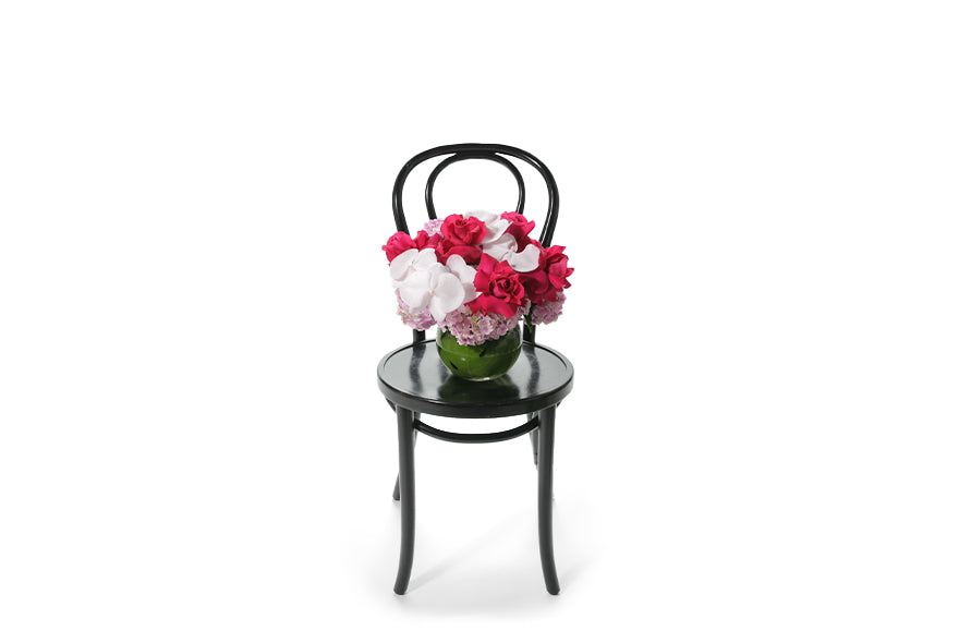 A pretty hot pink and blush pastel pink vase design sitting on a black bentwood chair with white background. 20cm ball vase, lined with green monsteria leaf displaying seasonal hot pink and blush pink flowers with no green foliage. Wide image of the vase design sitting on chair.
