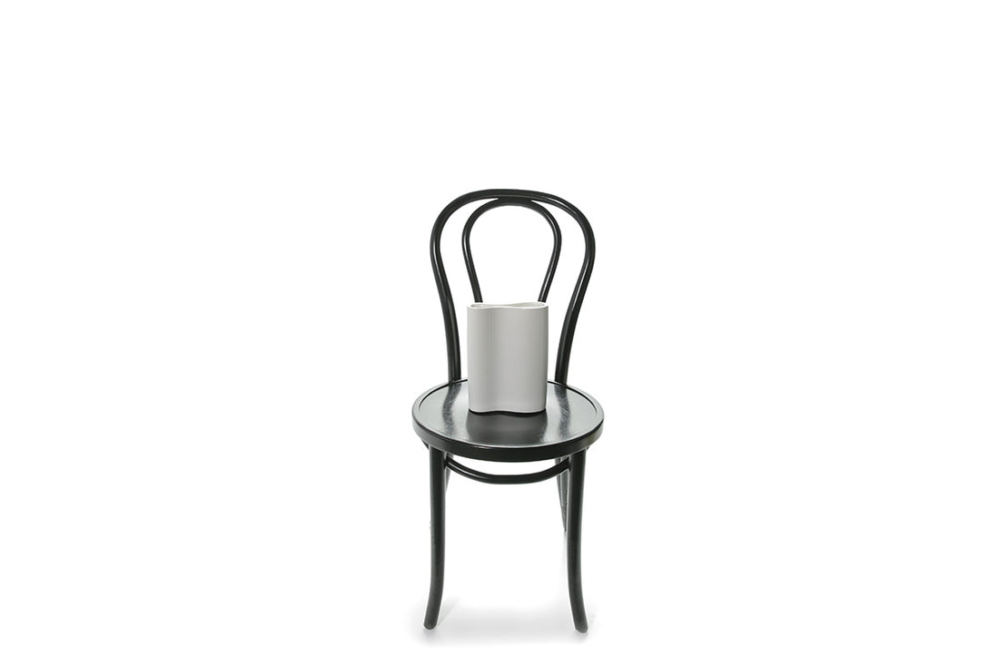 A 22cm White ceramic wave vase sitting on black bentwood chair with white background.