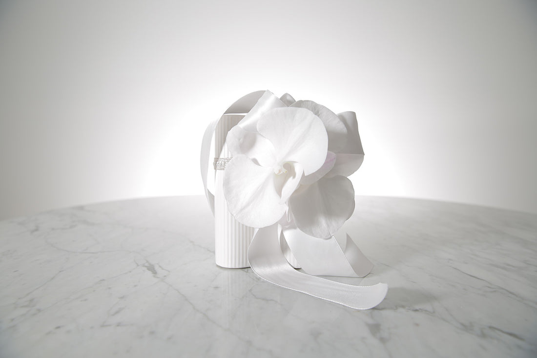 Kate Hill Flowers white phalaenopsis orchid wrist corsage with satin bows on a silver elastic wristlet, set on a marble table.