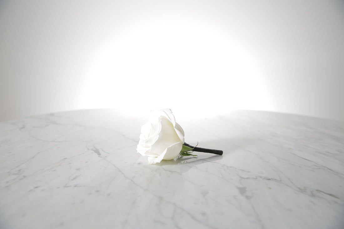 Kate Hill Flowers white rose buttonhole flower with black-taped stem and pearl pin, lapel-ready on a marble surface with a light grey background.