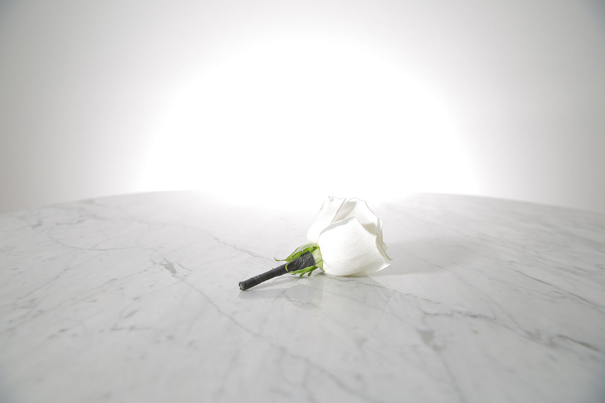Kate Hill Flowers white rose buttonhole flower with black-taped stem and pearl pin, lapel-ready on a marble surface with a light grey background.