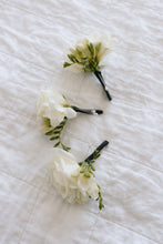 3 Buttonholes made with white freesias and black stems, laying on white linen bed. 