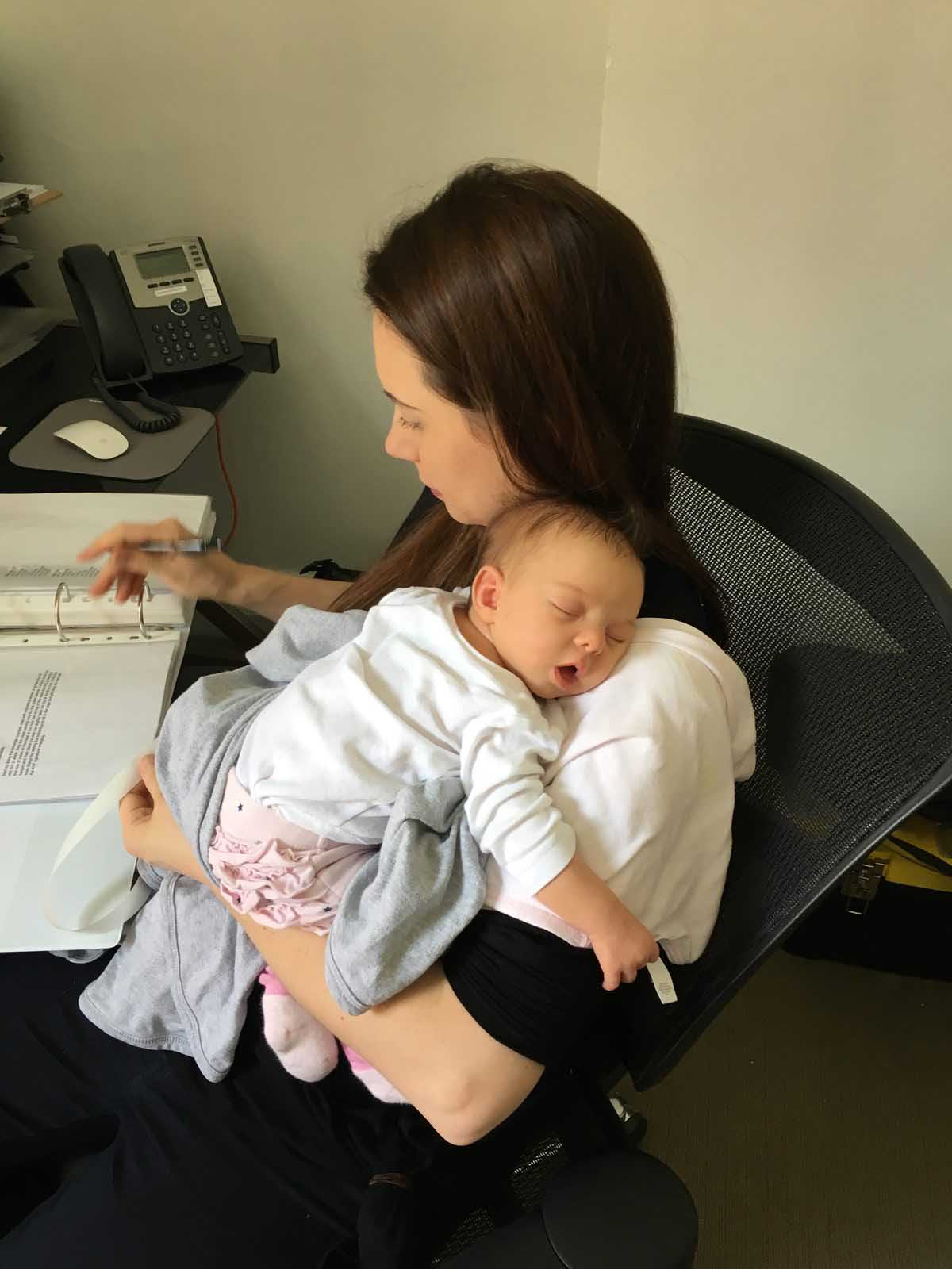 Kate Hill working at her studio desk with her newborn daughter asleep on her chest