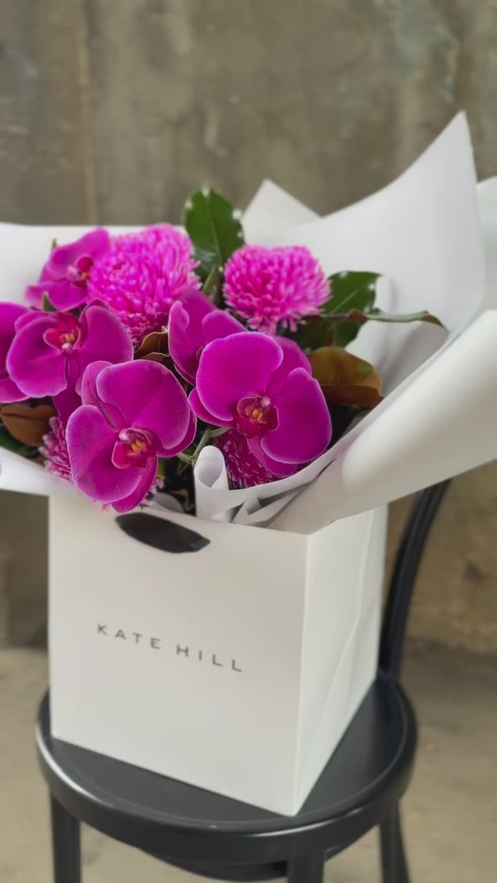 Video of a vibrant hot pink and magenta flower bouquet, presented in our signature white wrapping and flower bag. Bouquet bag is sitting on a black bentwood chair with a concrete background.