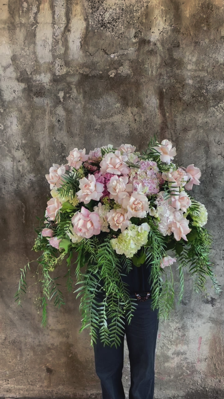 Video of Classic blush roses, blush hydrangeas, lilac stock and foliages feature in a Pastel pink, white and green casket design, held by a florist wearing back, up against a concrete wall. Flowers featuring blush classic roses, blush and white hydrangeas, peppercorn foliage. 