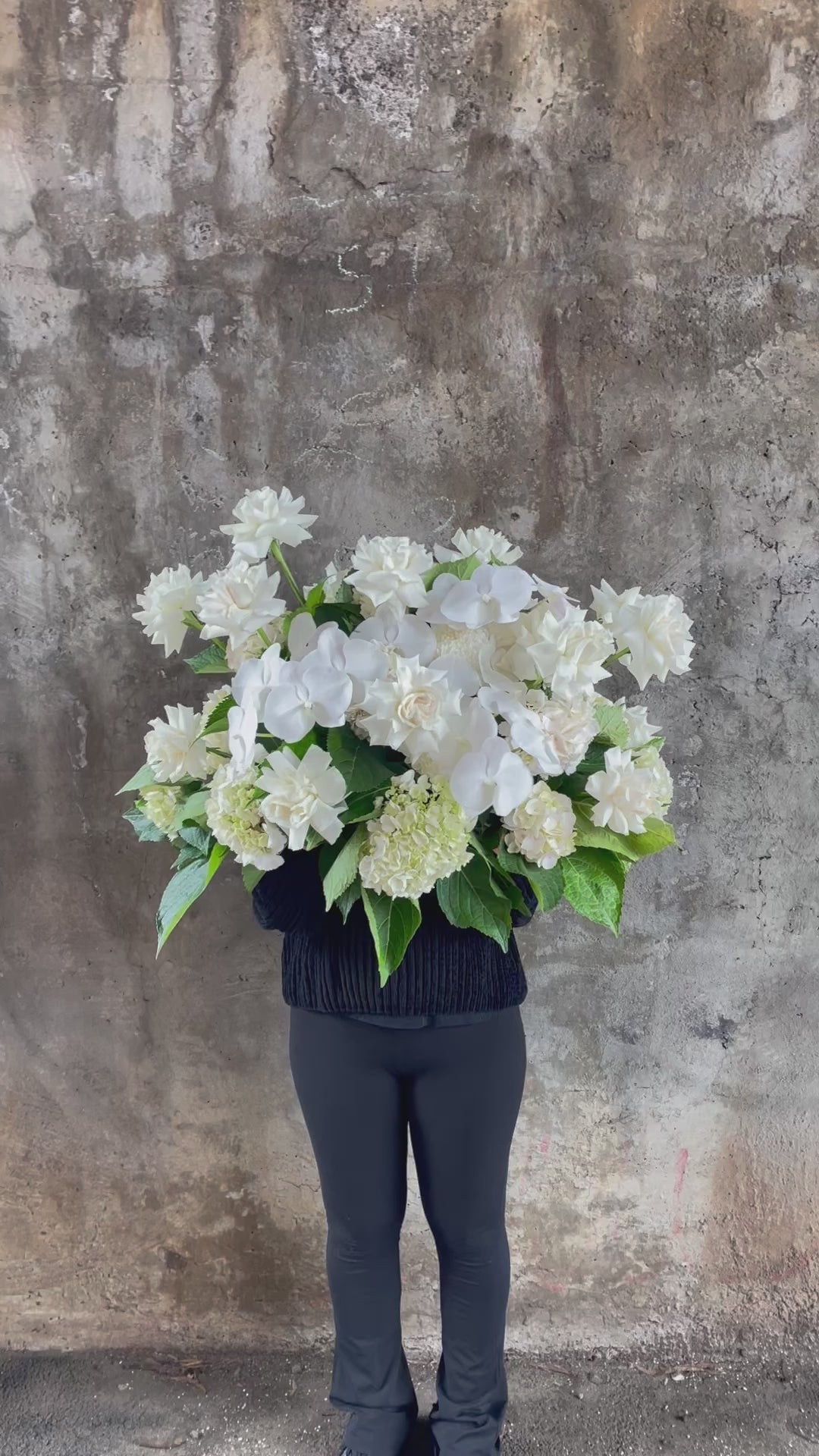 Video of Large white and green casket tribute design, being held by a florist wearing black and standing in front of a concrete wall.