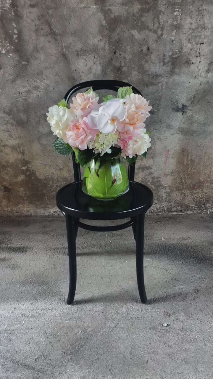 Video of Blush white and green flowers displayed in a leaf lined tapered vase, sitting on a black bentwood chair with a concrete wall background.