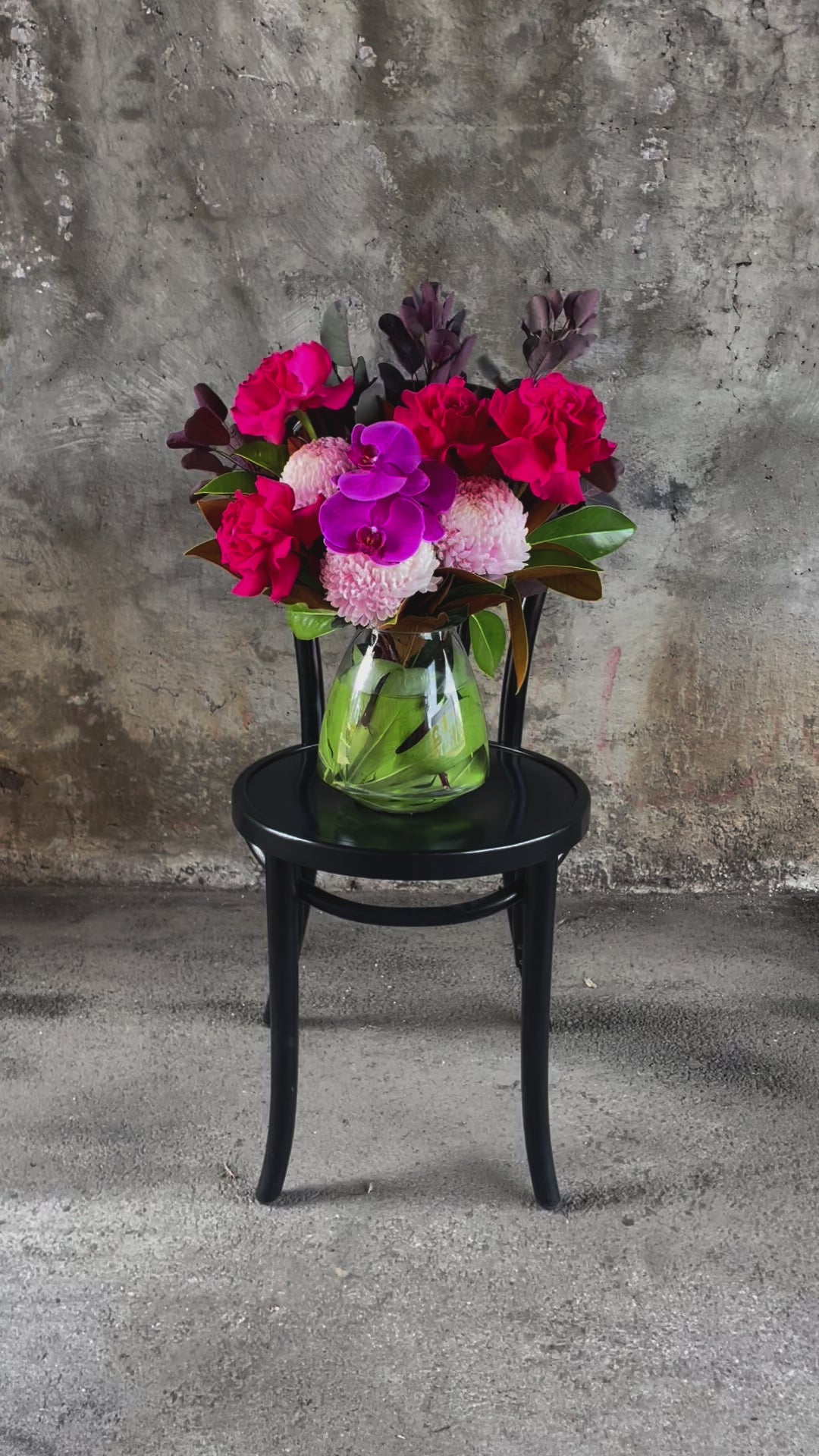 Video of Hot pink, blush, magenta flowers and seasonal foliage displayed in a leaf lined tapered vase, sitting on a black bentwood chair with a concrete wall background.