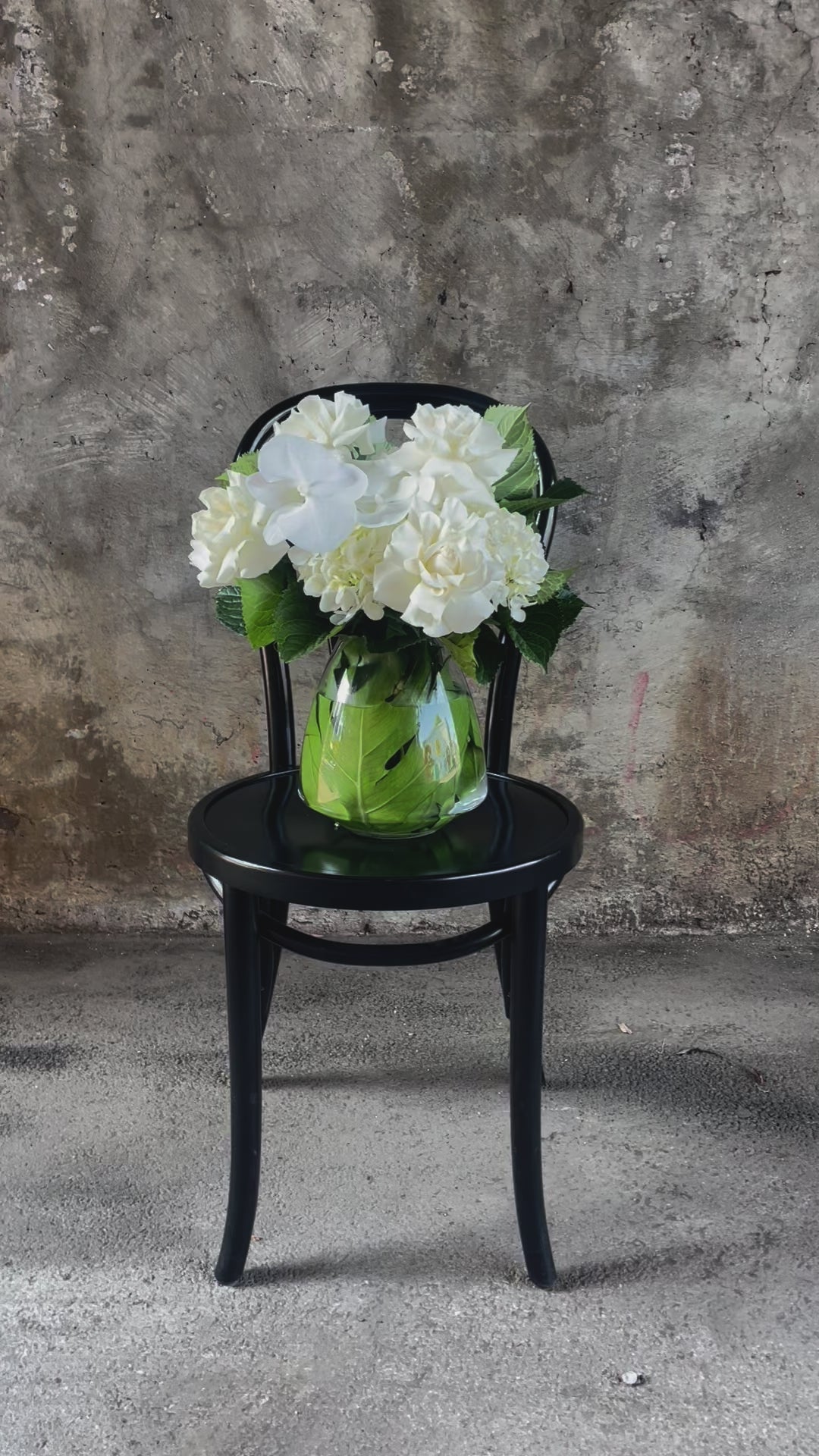 Video of White and green flowers displayed in a glass tapered vase, sitting on a black bentwood chair with a concrete wall background.