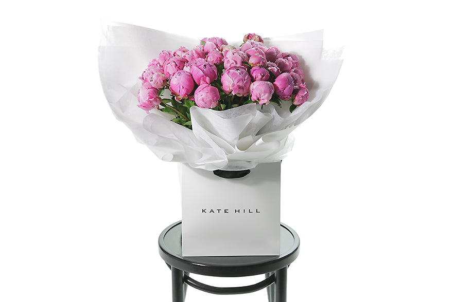 Best selling Peony Rose bouquet featuring 15 stems of pink peonies (flower only). Bouquet of peony roses presented in white signature wrapping and placed into Kate Hill Flower Bag. Bouquet bag sitting on a black bentwood chair.