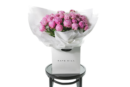 Best selling Peony Rose bouquet featuring 15 stems of pink peonies (flower only). Bouquet of peony roses presented in white signature wrapping and placed into Kate Hill Flower Bag. Bouquet bag sitting on a black bentwood chair.