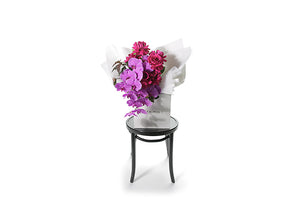 Wide image of the large bouquet to give you a honest perspective of the bouquet size. Chic, large and luxe bouquet featuring magenta phalaenopsis orchid stems, hot pink reflexed rose stems and seasonal burgundy foliages. Beautifully wrapped in our signature white wrapping.Gift bouquet presented in Kate Hill Flower Bag. Large and luxurious gift flower bouquet sitting on a black bentwood chair.