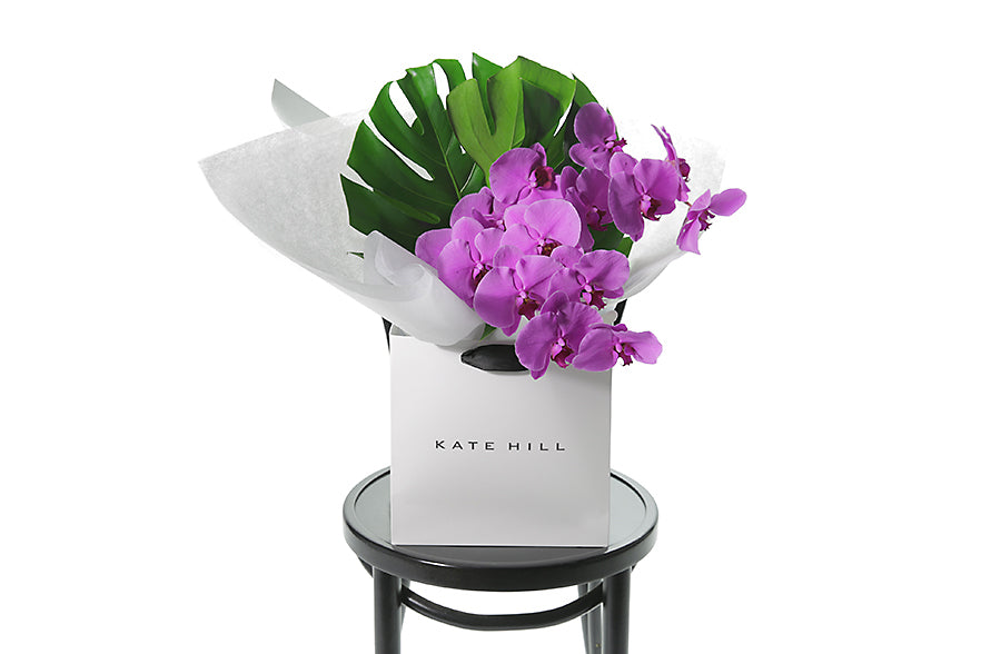 A vibrant and luxe flower bouquet featuring 2 magenta phalaenopsis orchid stems and lush green monsteria leaves. The bouquet is wrapped beautifully in the Kate Hill Flowers signature style with white wrapping and placed into the flower bag to ensure the bouquet remains fresh. Bouquet bag is sitting on a black bentwood chair.