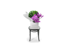 Chic and long lasting gift bouquet featuring 2 stems of Magenta phalaenopsis orchids and lush green monsteria leaves. Bouquet presented in Kate Hill Flower Bag. Wide image of the bouquet bag sitting on the black bentwood chair so you can see the size of the bouquet.