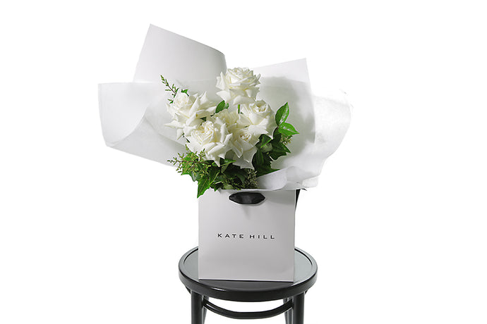 Large white rose bouquet displaying 6 stems of reflexed roses and green seasonal foliage. Presented beautifully in white signature wrapping and in Kate Hill Flower Bag. Flower bouquet bag sitting on a black bentwood chair.
