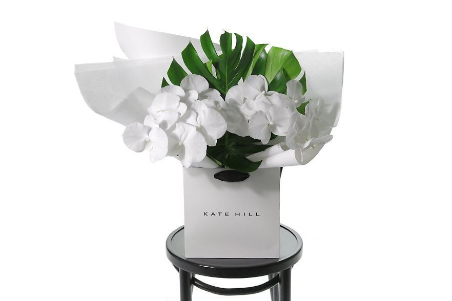 Chic and long lasting gift bouquet featuring 2 stems of pure white phalaenopsis orchids and lush green monsteria leaves. Bouquet presented in Kate Hill Flower Bag. Bouquet bag sitting on a black bentwood chair.